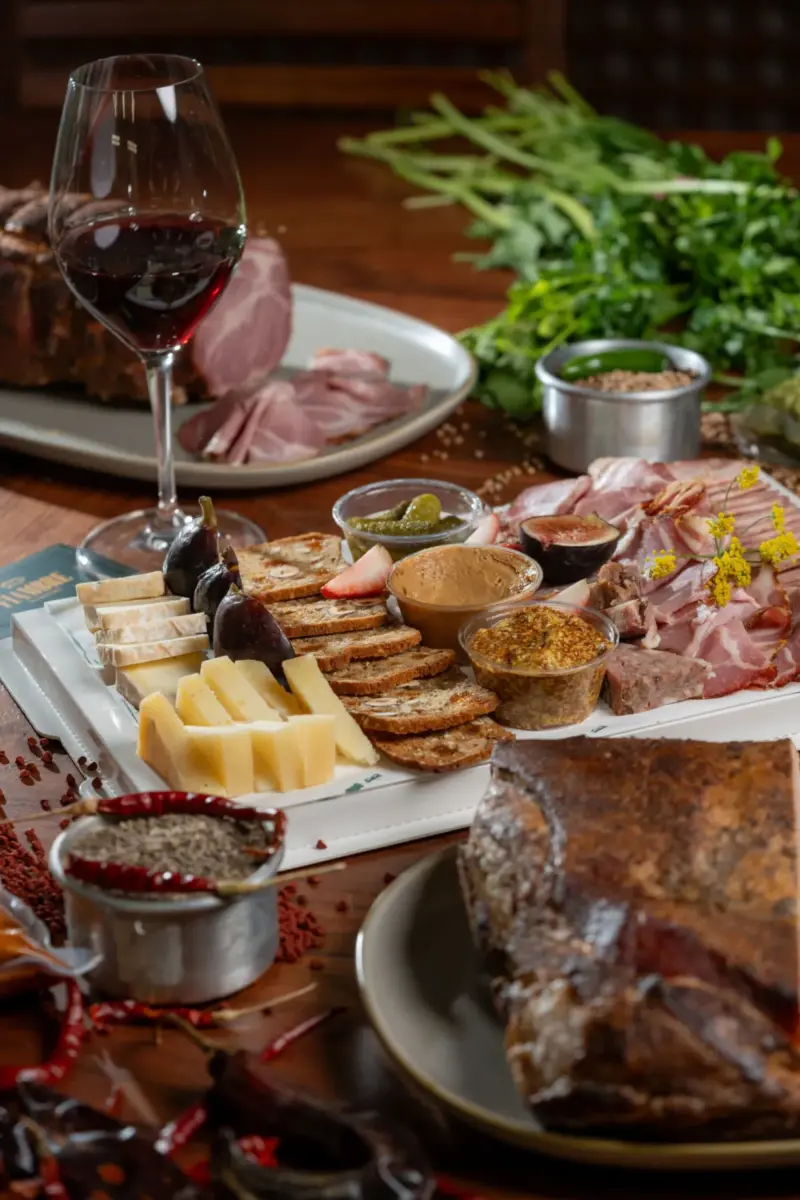 tabla de charcuteria con queso, pan y una copa de vino tinto de fondo