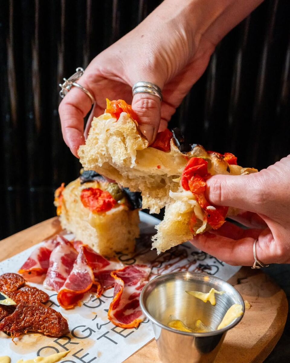 persona partiendo una focaccia con las manos, con una tabla de charcutería de fondo
