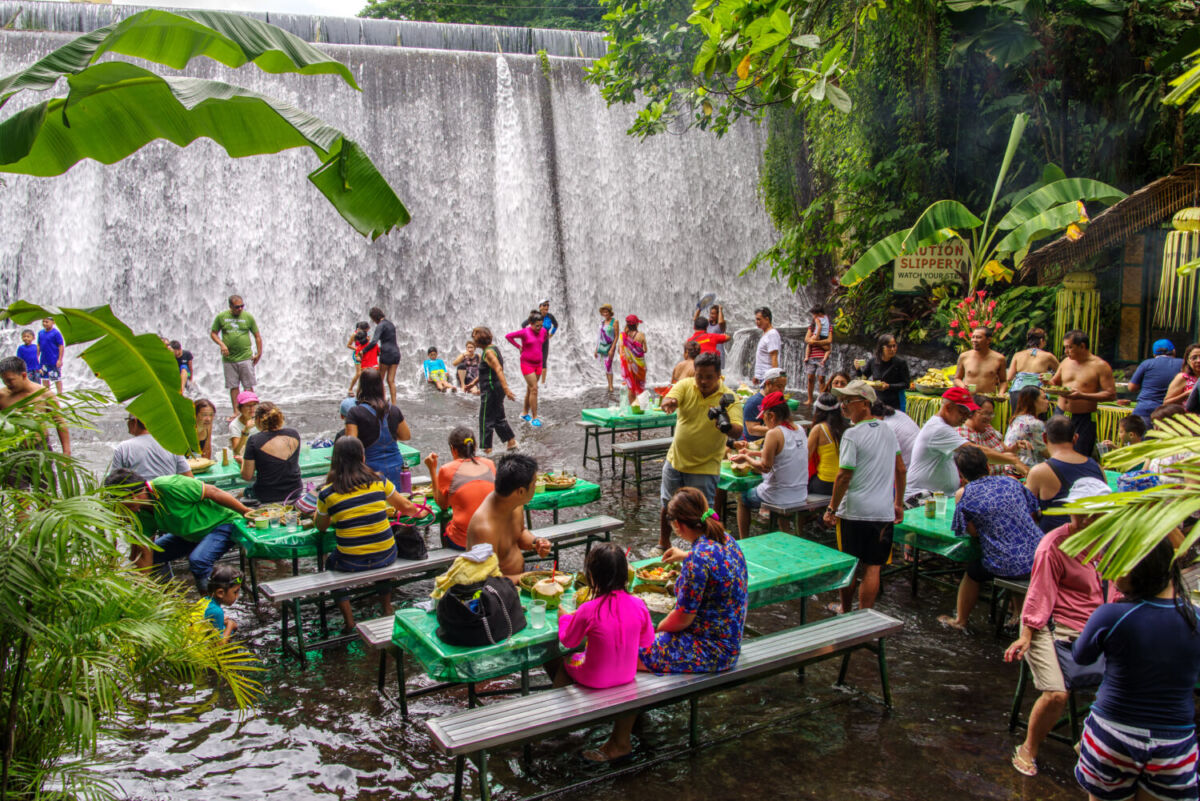 Gente comiendo sin zapatos y mesas sobre el agua en el único restaurante dentro de una cascada 