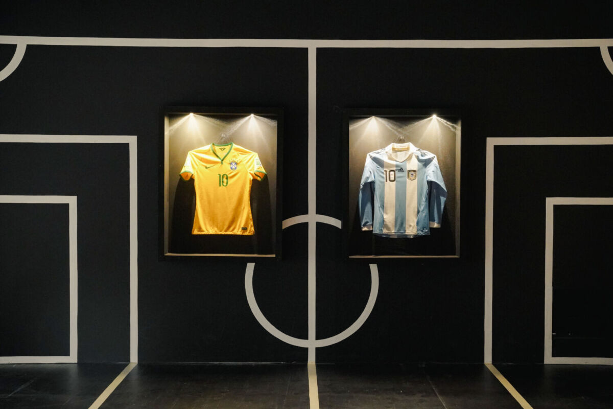 camisetas de futbol de Maradona y Pelé en dos vitrinas iluminadas contra un fondo negro con las líneas blancas de una cancha de futbol