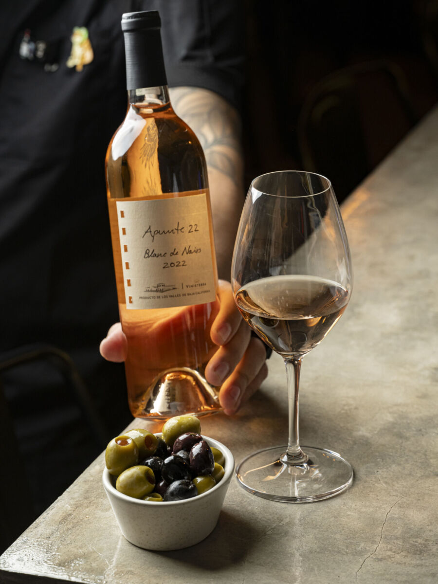 Botella de vino con una copa servida a un lado y un platón de aceitunas 