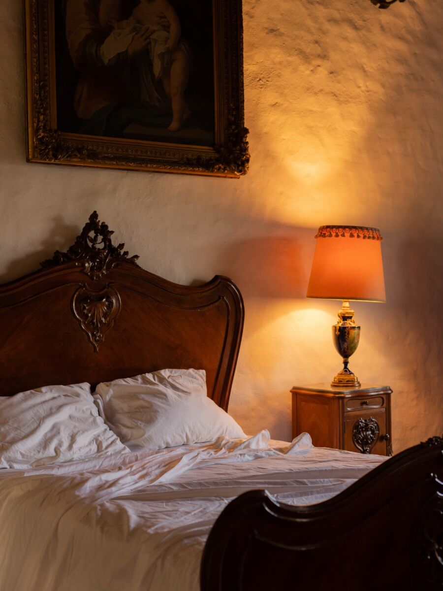 close up de una cama con sábanas blancas, cabecera de madera tallada y una lámpara de luz cálida encendida sobre el buró