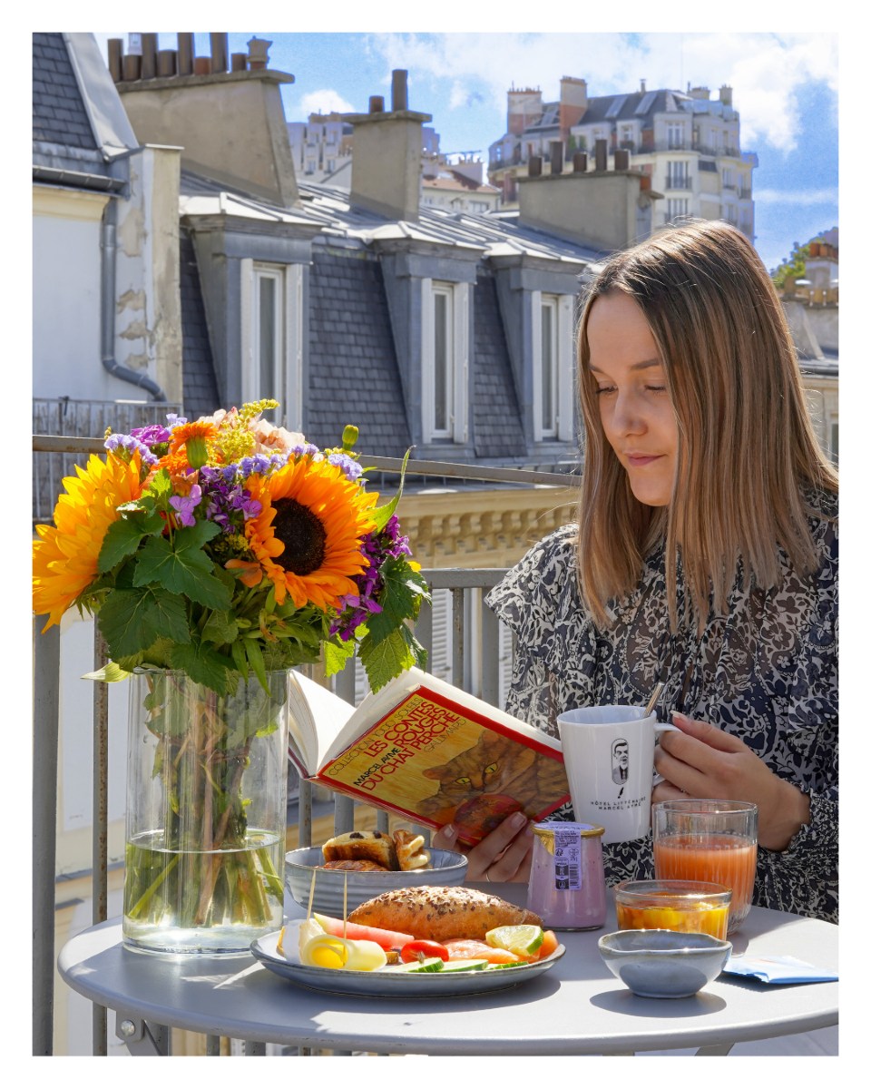 mujer rubia joven leyendo un libro mientras bebe café y desayuna fruta en una mesa con flores en un balcón con los techos parisinos de fondo