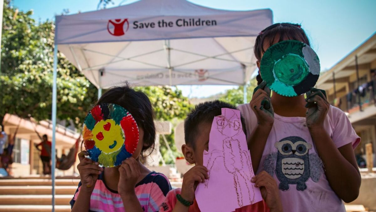 tres niños cubriendo sus rostros con máscaras hechas a mano frente a una carpa de Save the Children