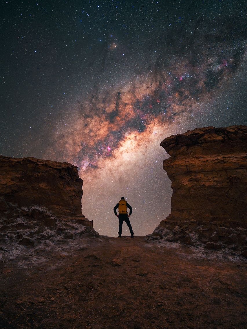 foto panoramica de una persona posando de espaldas entre dos formaciones rocosas con la vía láctea de fondo