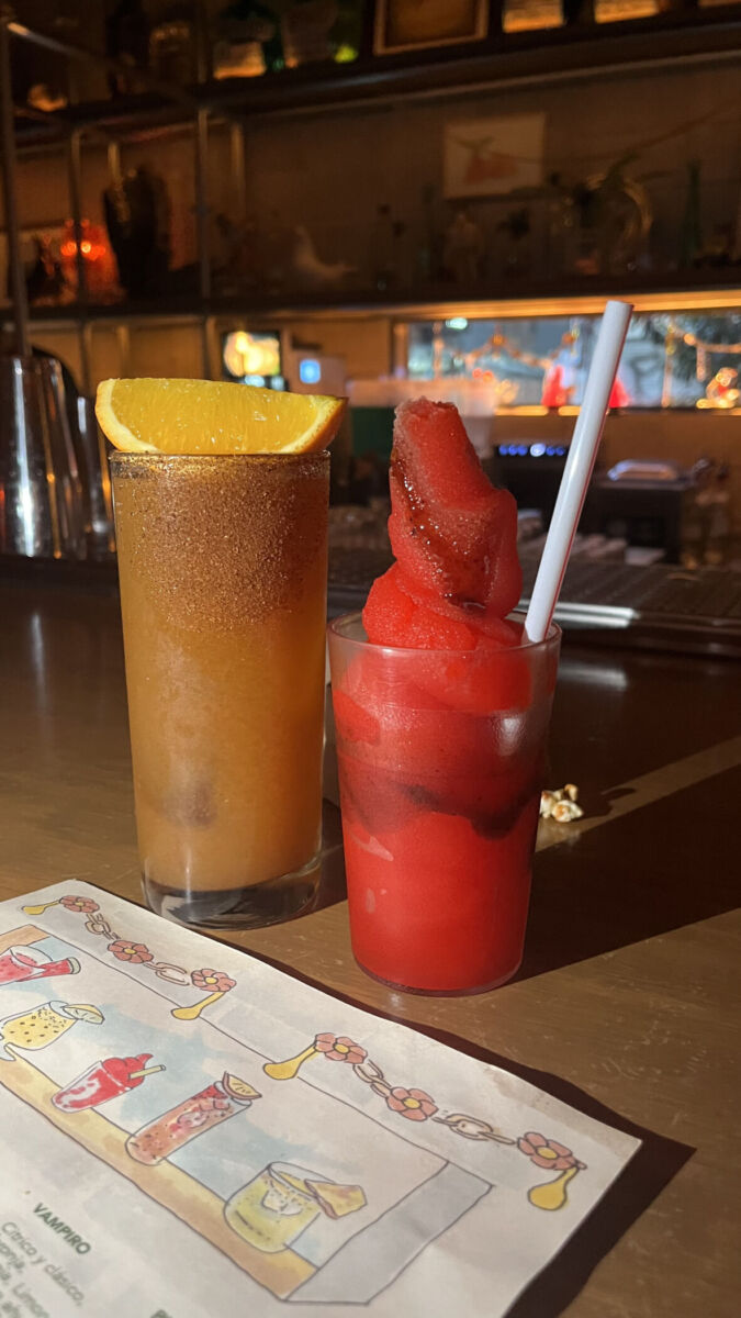 vaso escarchado con una bebida ámbar y un pedazo de naranja encima, junto a otro vaso con un slushy rojo con chile y popote