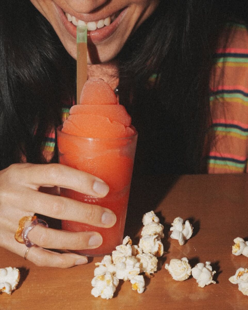 close up de mujer bebiedo un slushy de cereza con popote verde y palomitas en la mesa