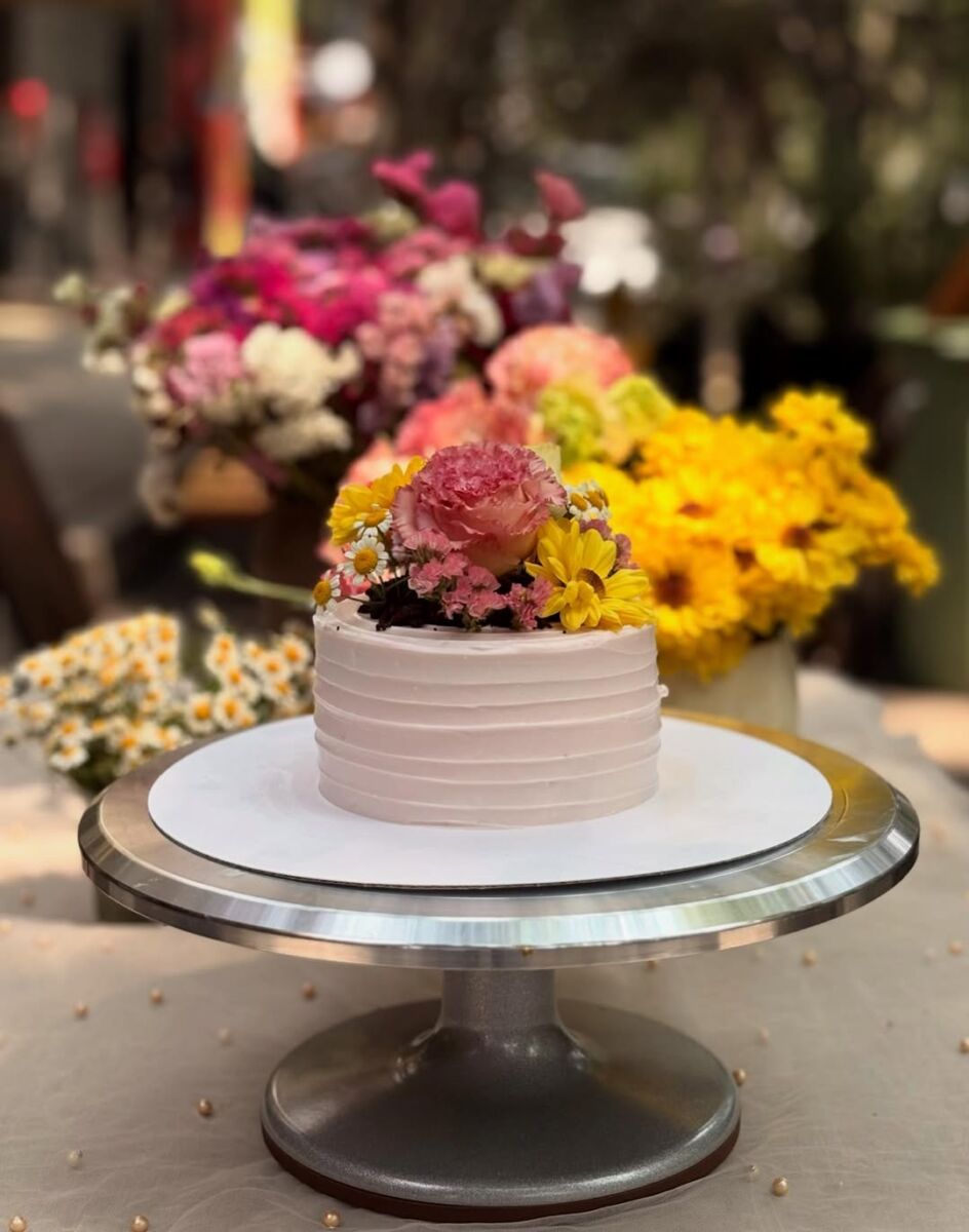 pastel individual con betún rosa cubierto de flores, con jarrones flores en el fondo