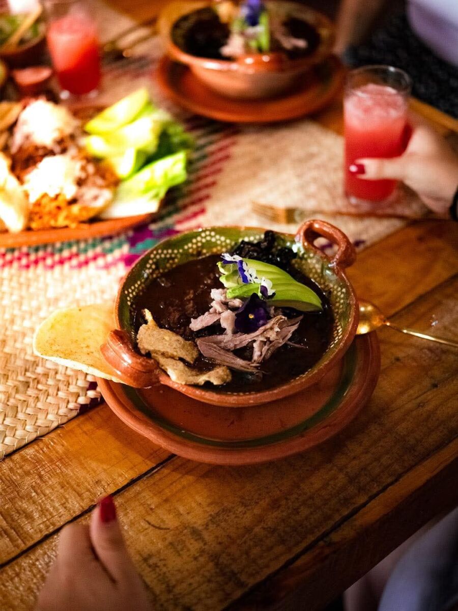 Pozole negro de Xochitepec servido en cazuela de barro con carne de cerdo, decorado con aguacate y flores pensamiento violetas, acompañado de tostadas y bebidas de sandía sobre mesa de madera rústica
