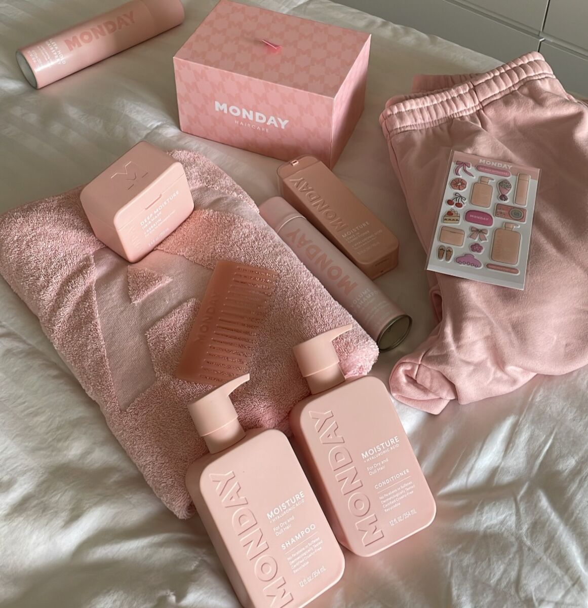 caja, pants, botellas, una toalla y stickers color rosa pastel colocados sobre una tela blanca