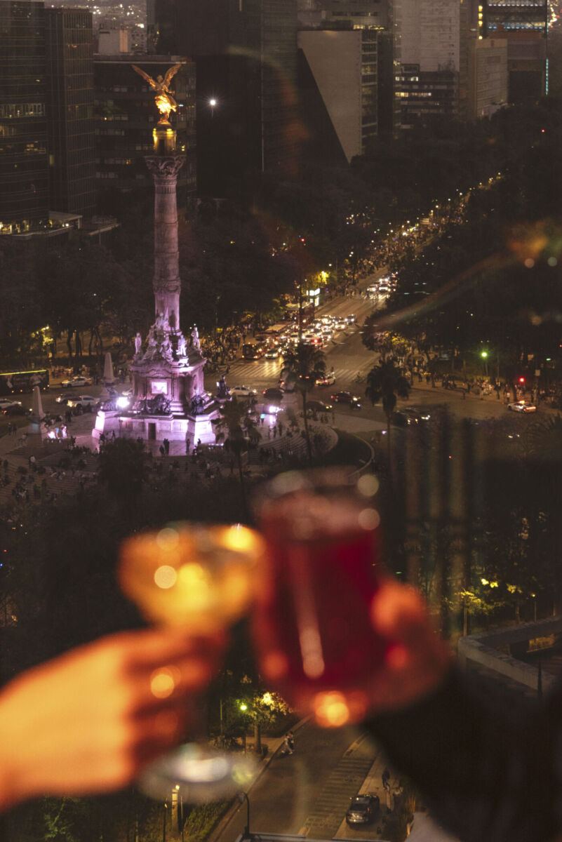 Dos personas brindando dentro de L’Obsidian Bar y al fondo el Ángel de la Independencia
