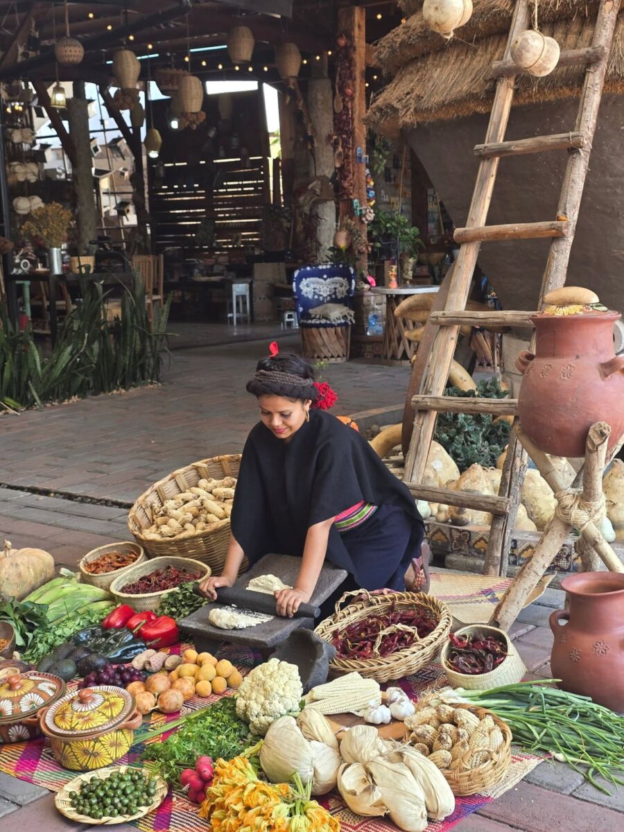 Cocinera tradicional trabajando entre canastos con maíz, chiles, verduras y utensilios artesanales dentro de Casa Mashihua.