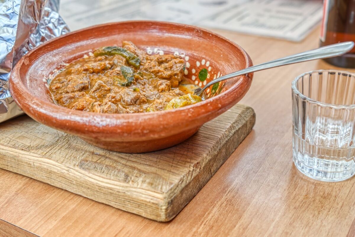 Cazuela de barro con guiso de carne y rajas de chile poblano, servida sobre una tabla de madera con un vaso de agua al lado, en un restaurante de Durango.