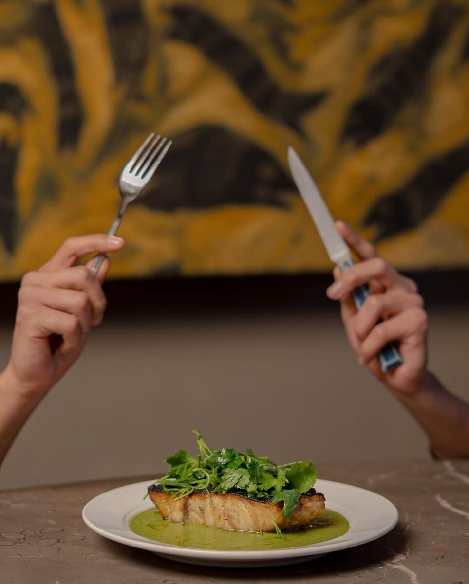 manos sosteniendo tenedor y cuchillo sobre un plato de filete de pescado con salsa verde y hierbas