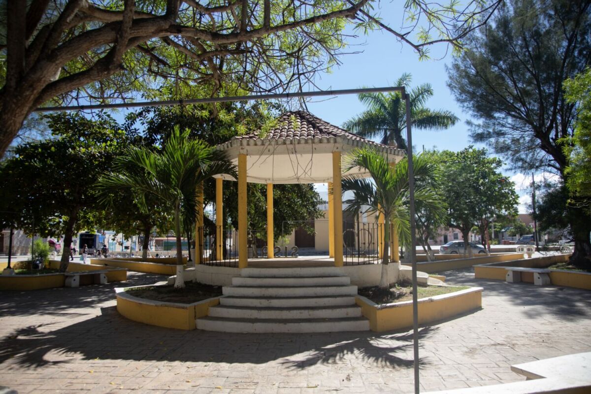 Kiosco amarillo y blanco con letras "Sisal" en la plaza principal del Pueblo Mágico, rodeado de palmeras y árboles tropicales, Yucatán