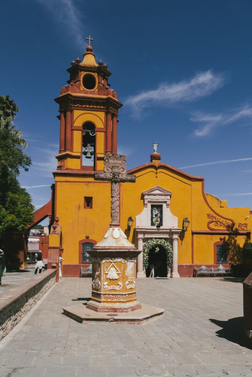 Cruz de piedra tallada frente a la fachada amarilla de una iglesia colonial en Bernal, Querétaro
