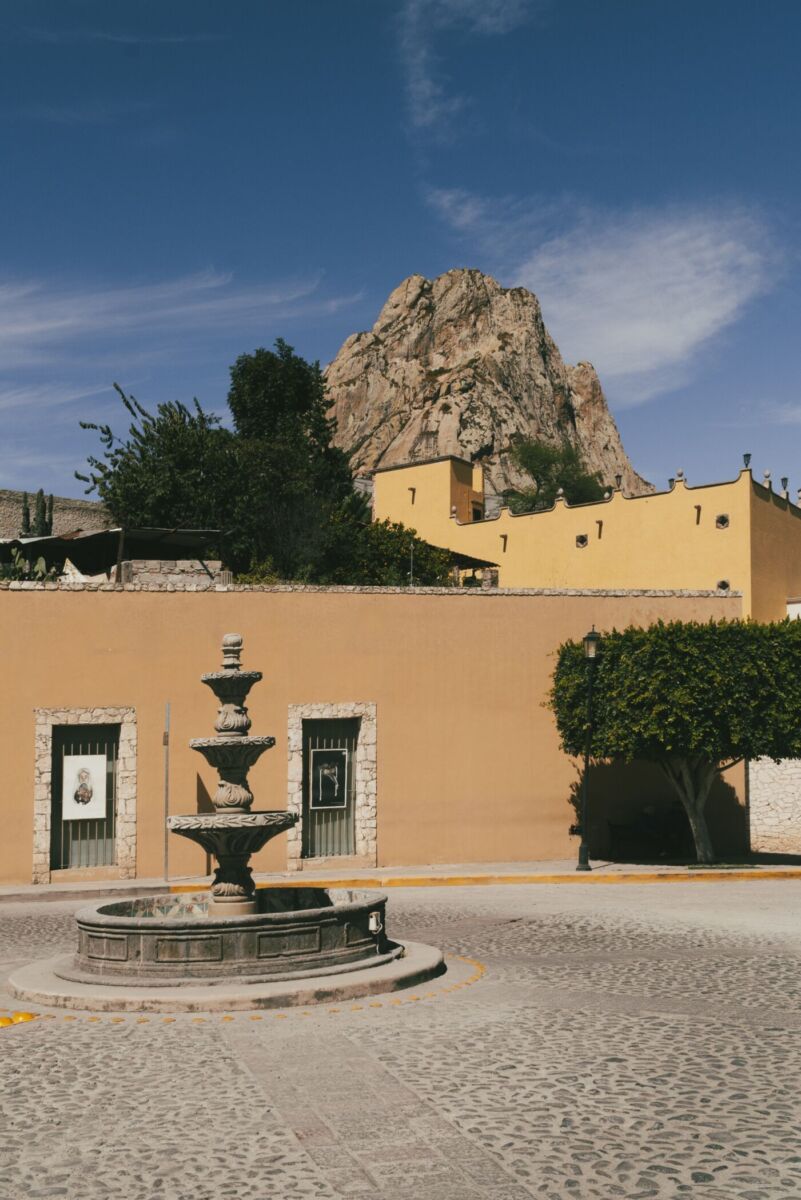 Fuente de piedra colonial con la Peña de Bernal elevándose sobre los tejados amarillos de Querétaro