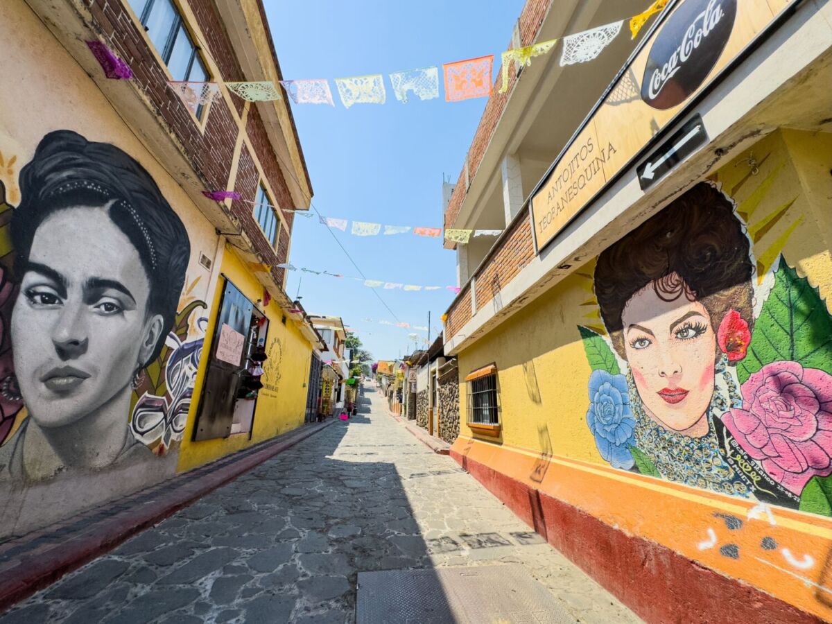 Callejón empedrado con murales de mujeres icónicas y papel picado colgando entre fachadas