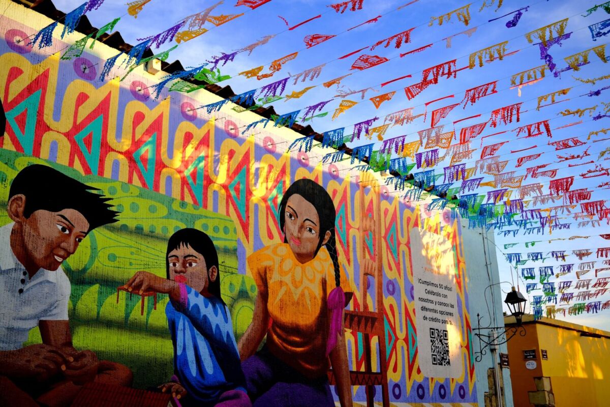 Mural colorido con niños indígenas bajo hileras de papel picado en un pueblo mexicano