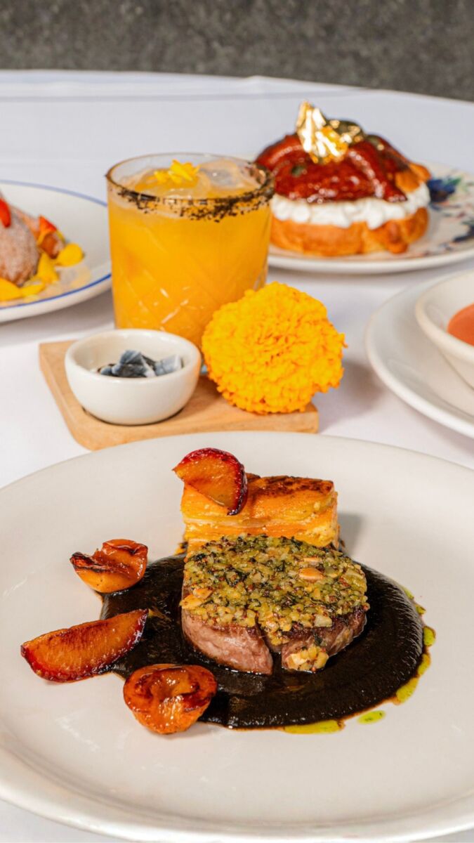 Pierna de cordero con costra de hierbas, sobre chichilo negro, con mil hojas de calabaza y ciruelas asadas, junto a un cóctel y un pan de muerto.