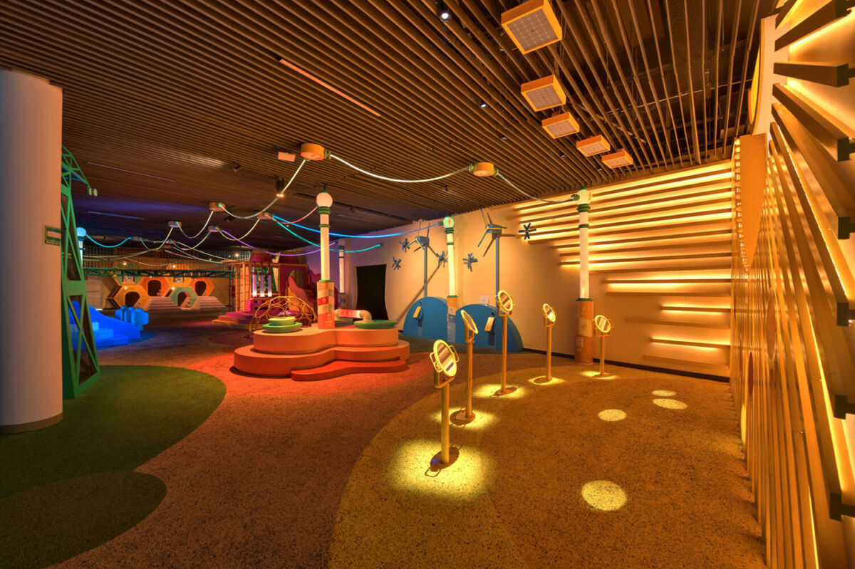 Sala de Niños MUNET con juegos de colores, lámparas amarillas sobre el piso y pasarelas de madera iluminadas en el techo.