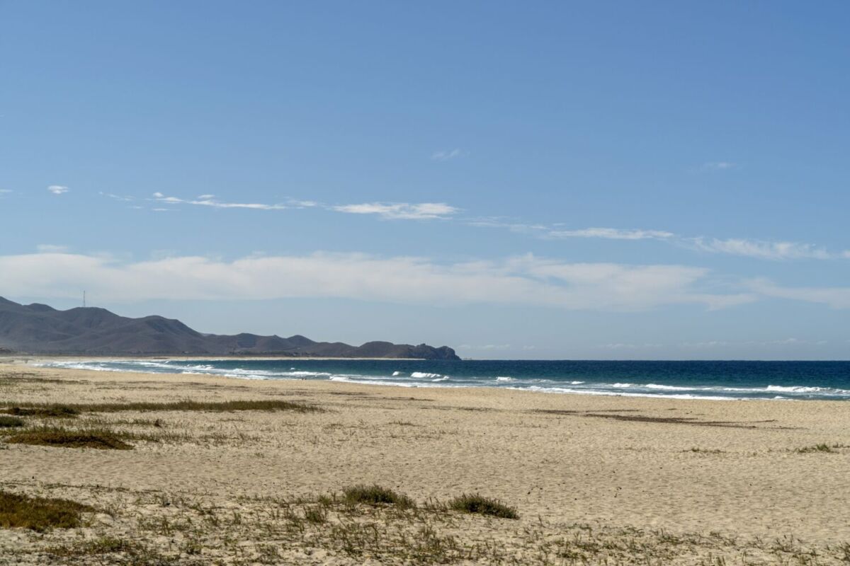 Playa de arena clara y vacía con olas azules rompiendo en la orilla y cerros secos al fondo, en Los Cerritos, Todos Santos
